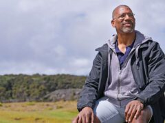 An African American man sits on a rock with hillside vista behind him.